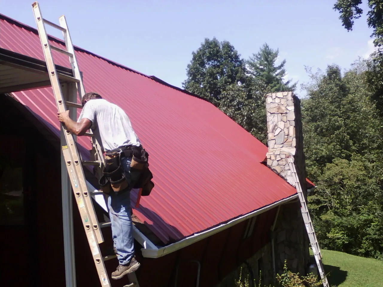 Roofer on a ladder beside a red metal roof and stone chimney — Soffit & Fascia Repair by Roofing Brothers in Lawrence